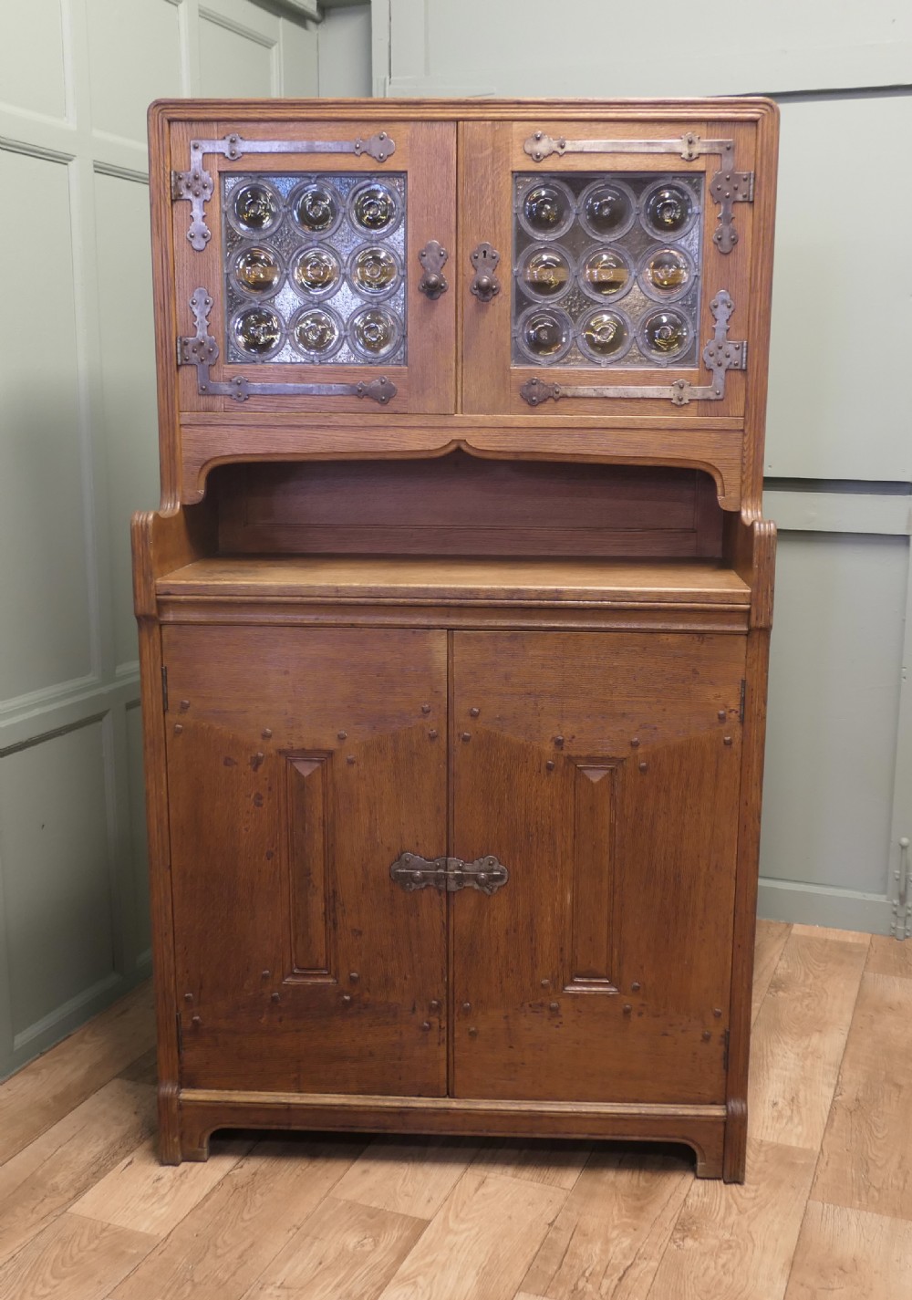A solid golden oak Arts and Crafts dresser featuring upper bullseye leaded glass doors with iron strap hinges above an open shelf and carved panelled lower doors.
