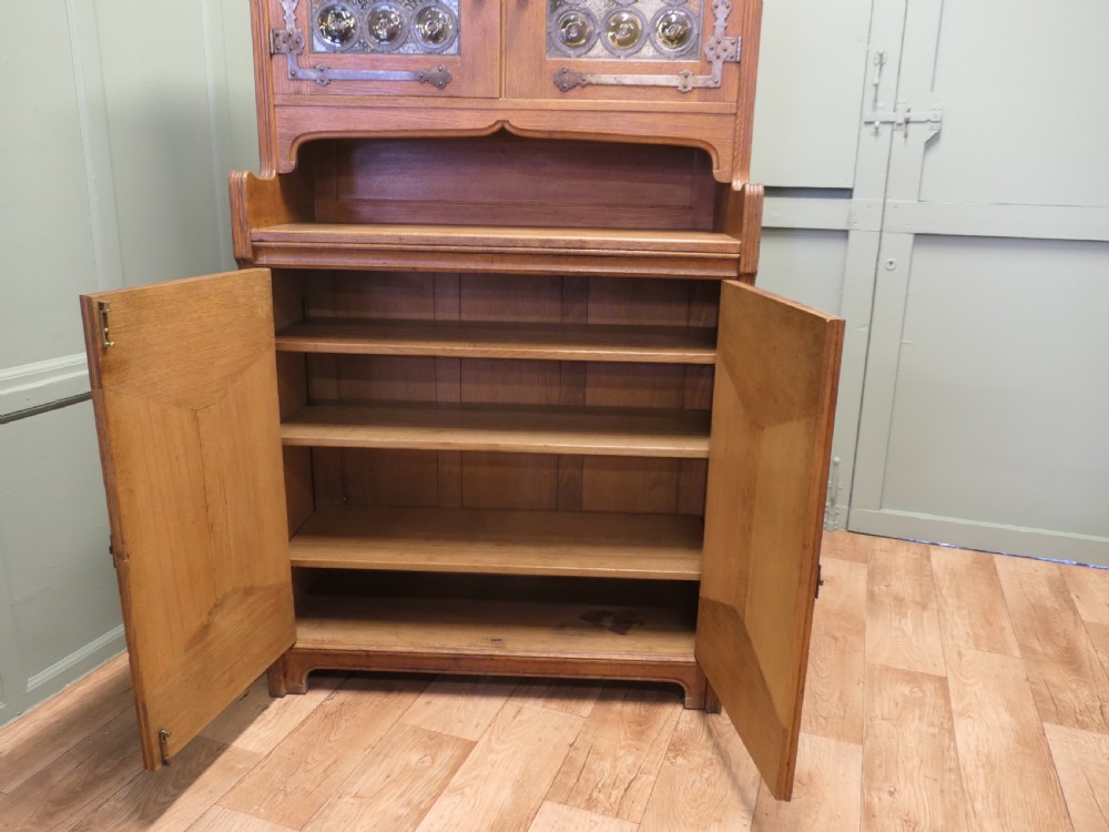 A solid golden oak Arts and Crafts dresser featuring upper bullseye leaded glass doors with iron strap hinges above an open shelf and carved panelled lower doors. View II