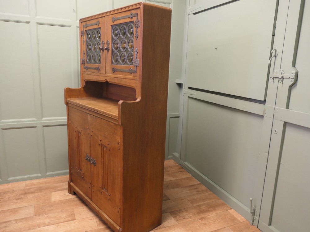 A solid golden oak Arts and Crafts dresser featuring upper bullseye leaded glass doors with iron strap hinges above an open shelf and carved panelled lower doors. View III