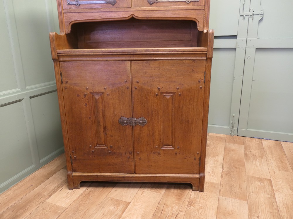 A solid golden oak Arts and Crafts dresser featuring upper bullseye leaded glass doors with iron strap hinges above an open shelf and carved panelled lower doors. View IV