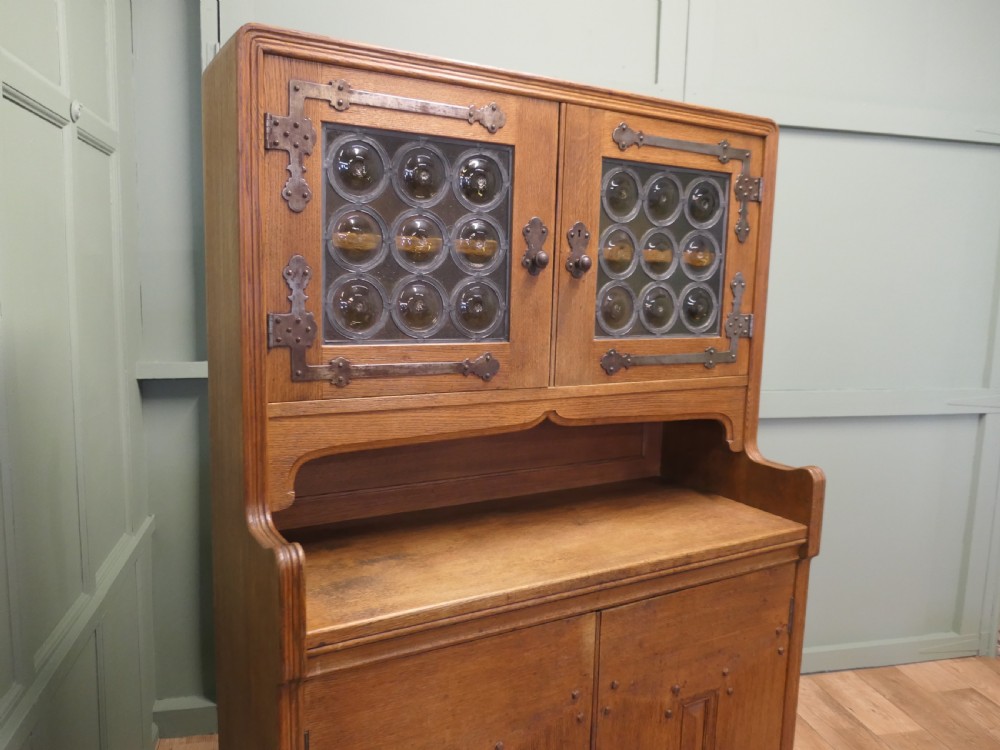 A solid golden oak Arts and Crafts dresser featuring upper bullseye leaded glass doors with iron strap hinges above an open shelf and carved panelled lower doors. View V