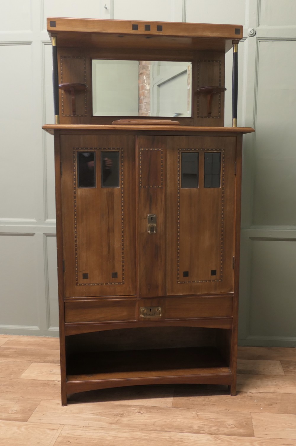 A Secessionist warm brown oak cocktail cabinet featuring a mirror-backed upper shelf supported by dark pillars, geometric wood inlay detailing on double doors, brass keyhole fittings, and an open lower shelf gallery.