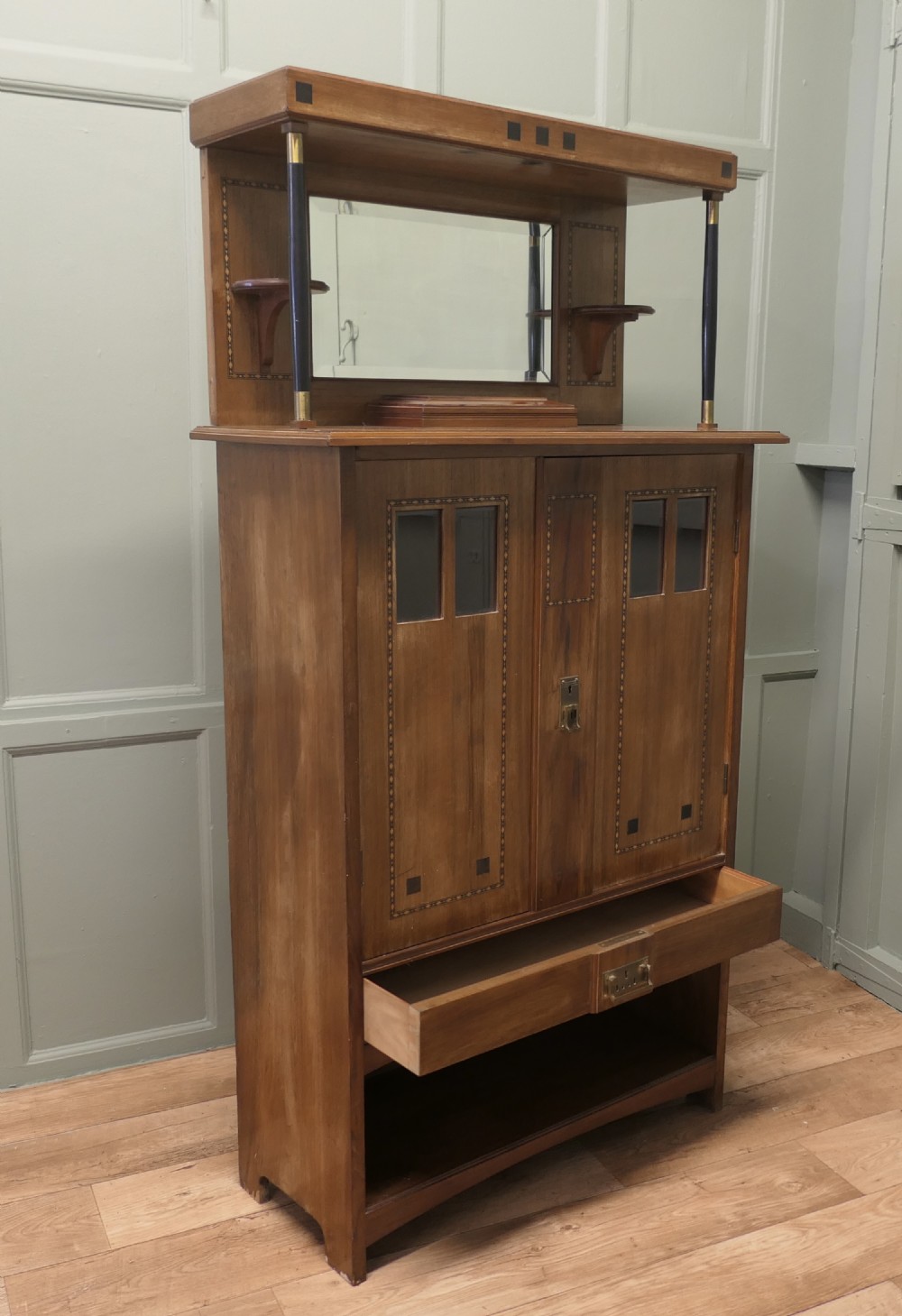 A Secessionist warm brown oak cocktail cabinet featuring a mirror-backed upper shelf supported by dark pillars, geometric wood inlay detailing on double doors, brass keyhole fittings, and an open lower shelf gallery. View II