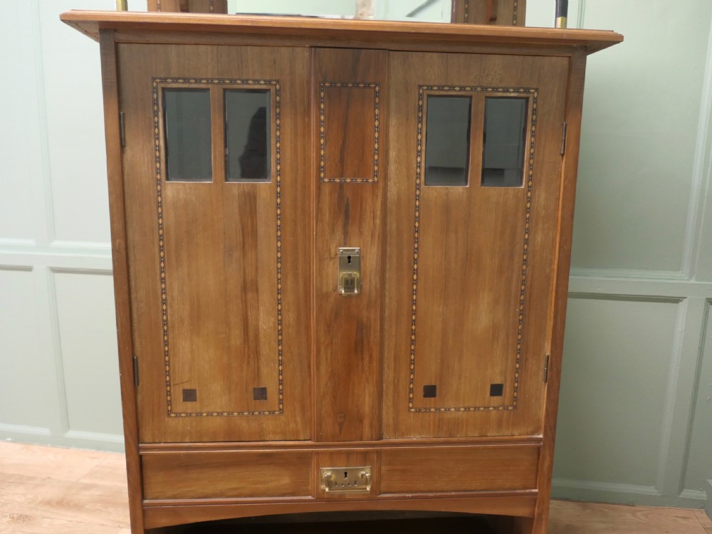 A Secessionist warm brown oak cocktail cabinet featuring a mirror-backed upper shelf supported by dark pillars, geometric wood inlay detailing on double doors, brass keyhole fittings, and an open lower shelf gallery. View III