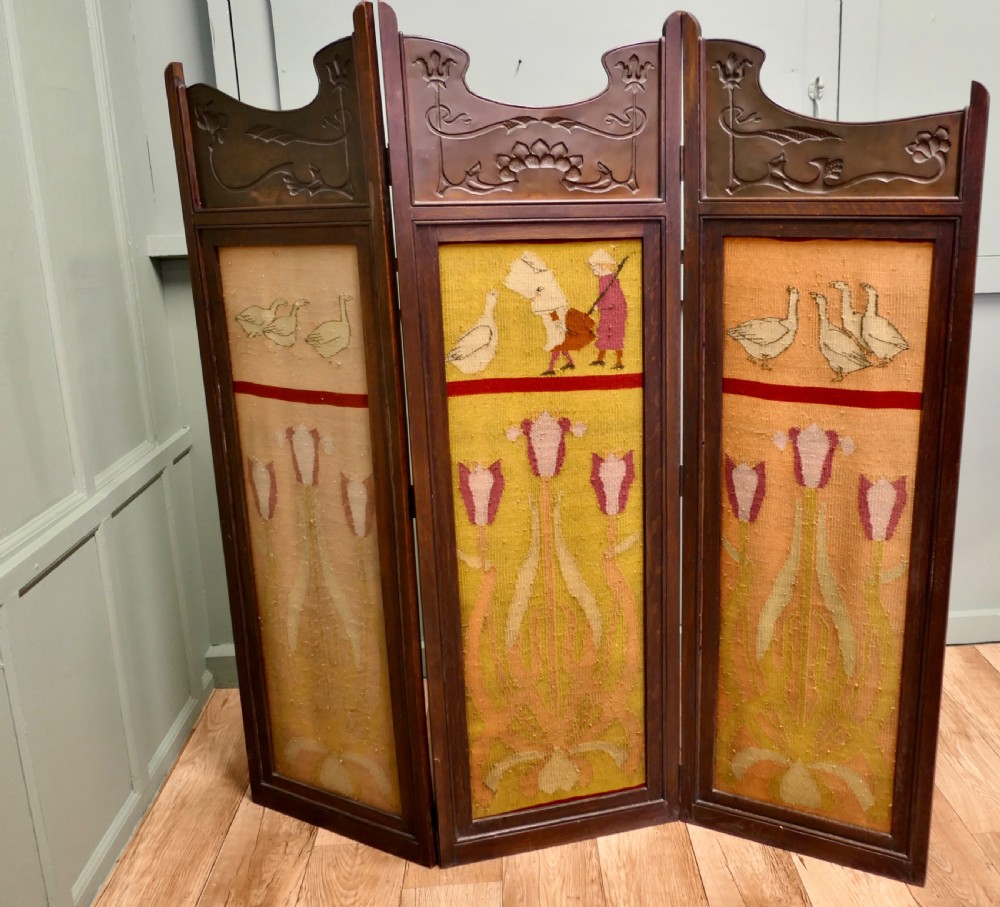 An Art Nouveau three-fold wooden room divider featuring carved oak frames, repousse copper top panels with stylised motifs, and woven wool textile sections depicting geese and floral designs.