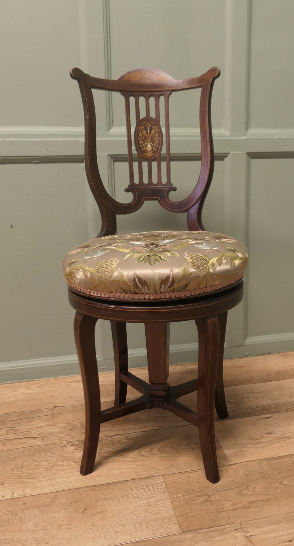 An antique mahogany revolving music chair featuring a lyre-shaped backrest with inlaid urn motif, a round embroidered seat cushion, and four splayed legs with a central cross-stretcher.