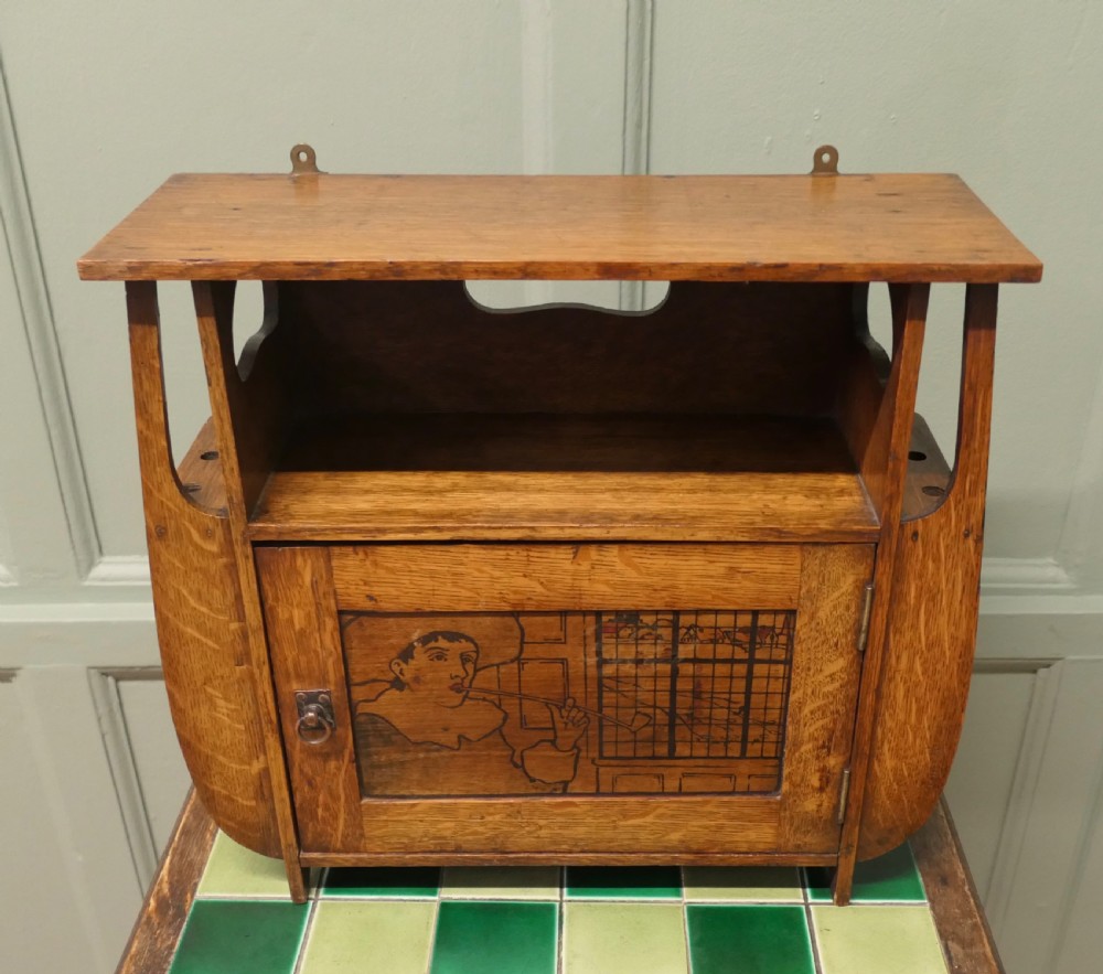 An Arts and Crafts oak wall cabinet featuring a single door decorated with a pyrography panel of a seated figure, framed by shaped side supports and a top shelf. View III