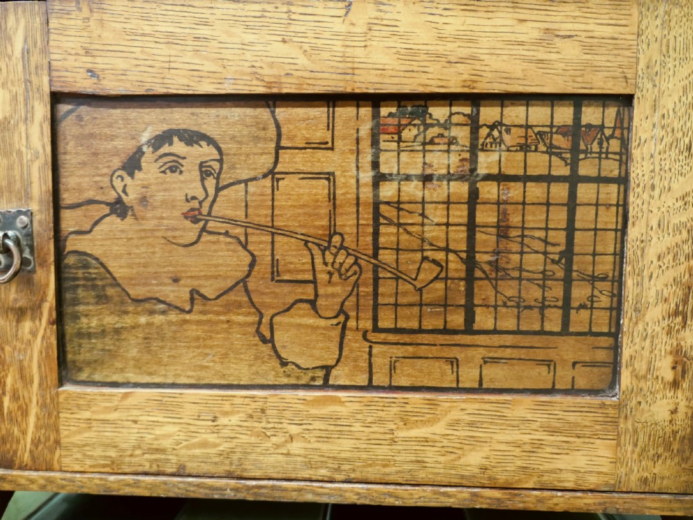 An Arts and Crafts oak wall cabinet featuring a single door decorated with a pyrography panel of a seated figure, framed by shaped side supports and a top shelf. View V
