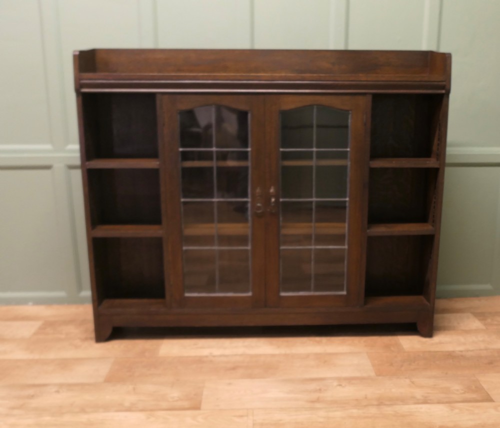 An Arts and Crafts dark oak bookcase featuring central leaded glass double doors flanked by open tier side shelves and a raised top gallery. View II