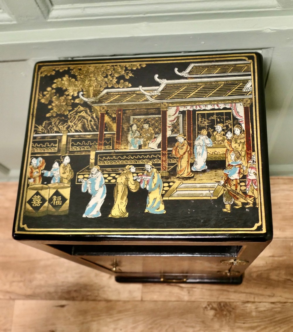 A narrow black lacquered Chinoiserie side cupboard featuring an open upper shelf, a hand-painted door with figures, and a bottom drawer with brass hardware. View II