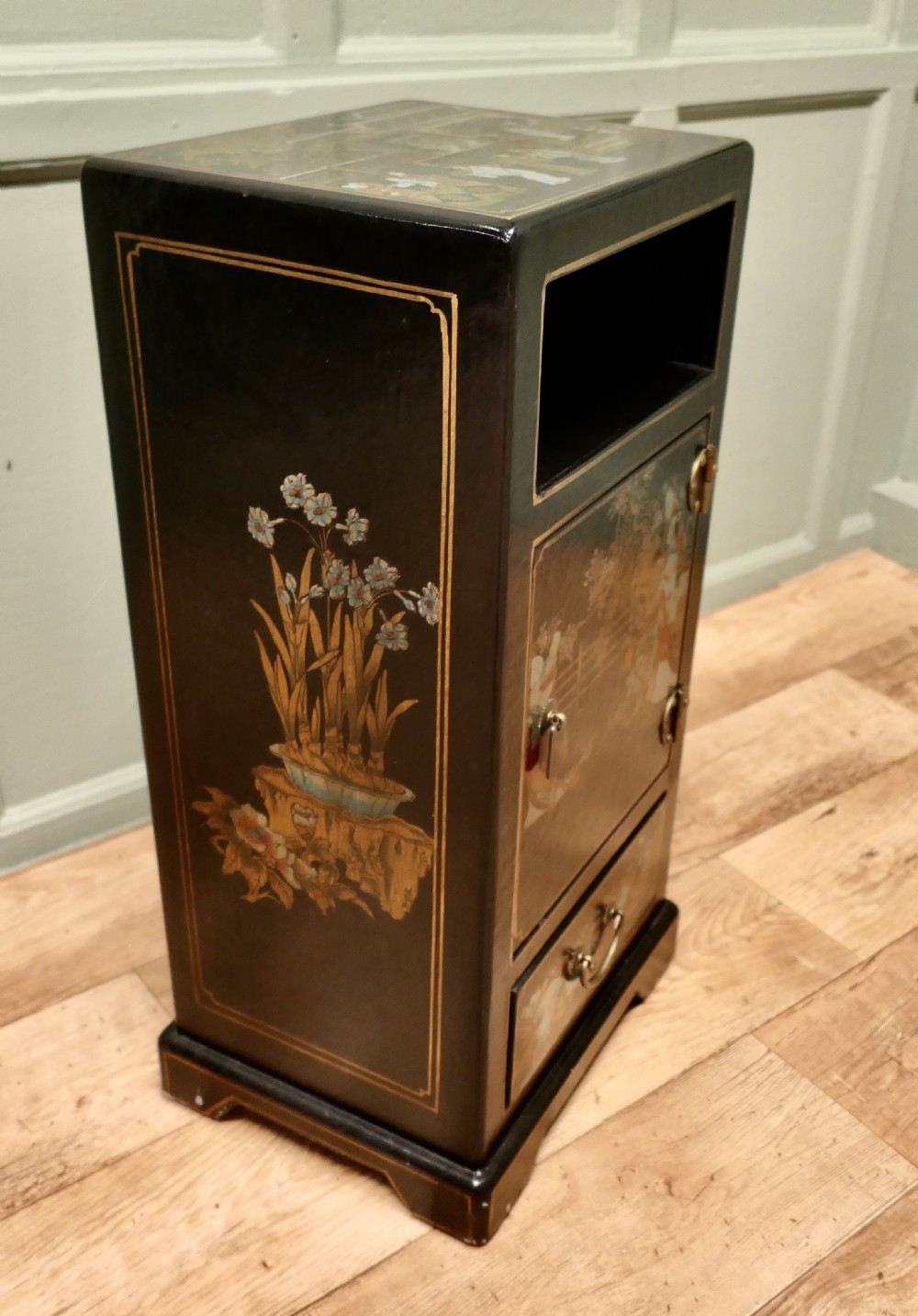 A narrow black lacquered Chinoiserie side cupboard featuring an open upper shelf, a hand-painted door with figures, and a bottom drawer with brass hardware. View V