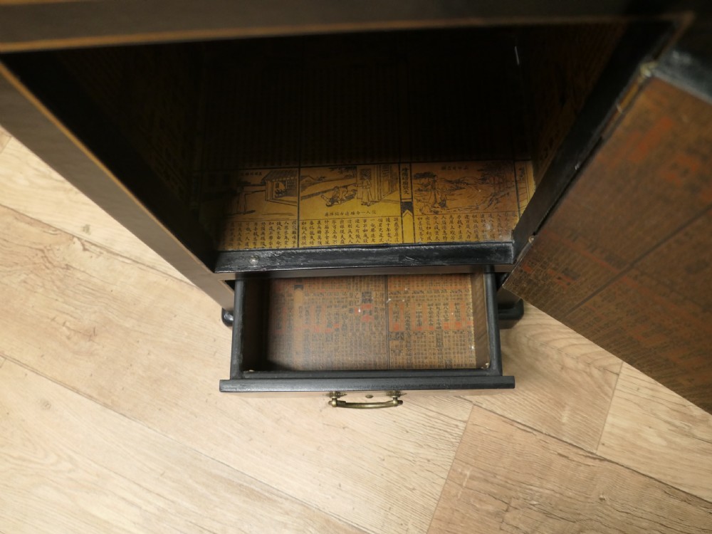 A narrow black lacquered Chinoiserie side cupboard featuring an open upper shelf, a hand-painted door with figures, and a bottom drawer with brass hardware. View VI