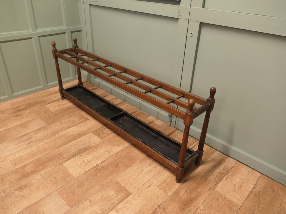 A six-foot long Arts and Crafts wooden umbrella stand with slatted compartment dividers, turned finials, and a removable metal drip tray base. View VI
