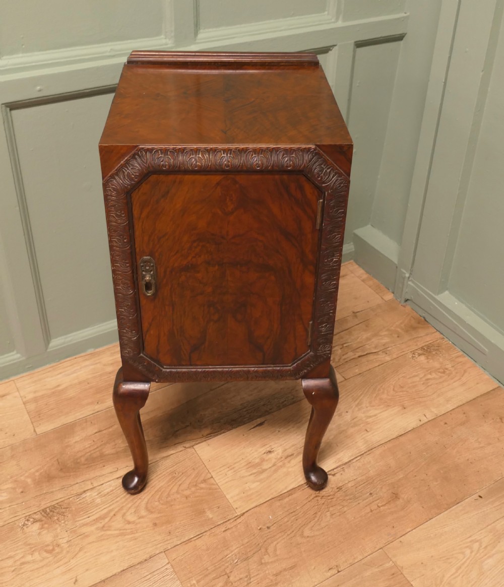An Art Deco burr walnut bedside cabinet featuring a carved door frame, brass latch, low gallery back, and four cabriole legs.