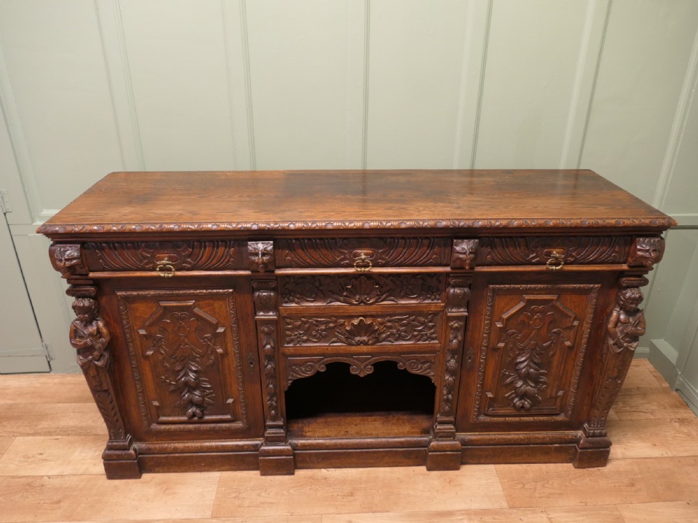 A heavily carved warm brown oak sideboard with three top drawers, relief carved cupboard doors, putti side supports, and a central open recess.