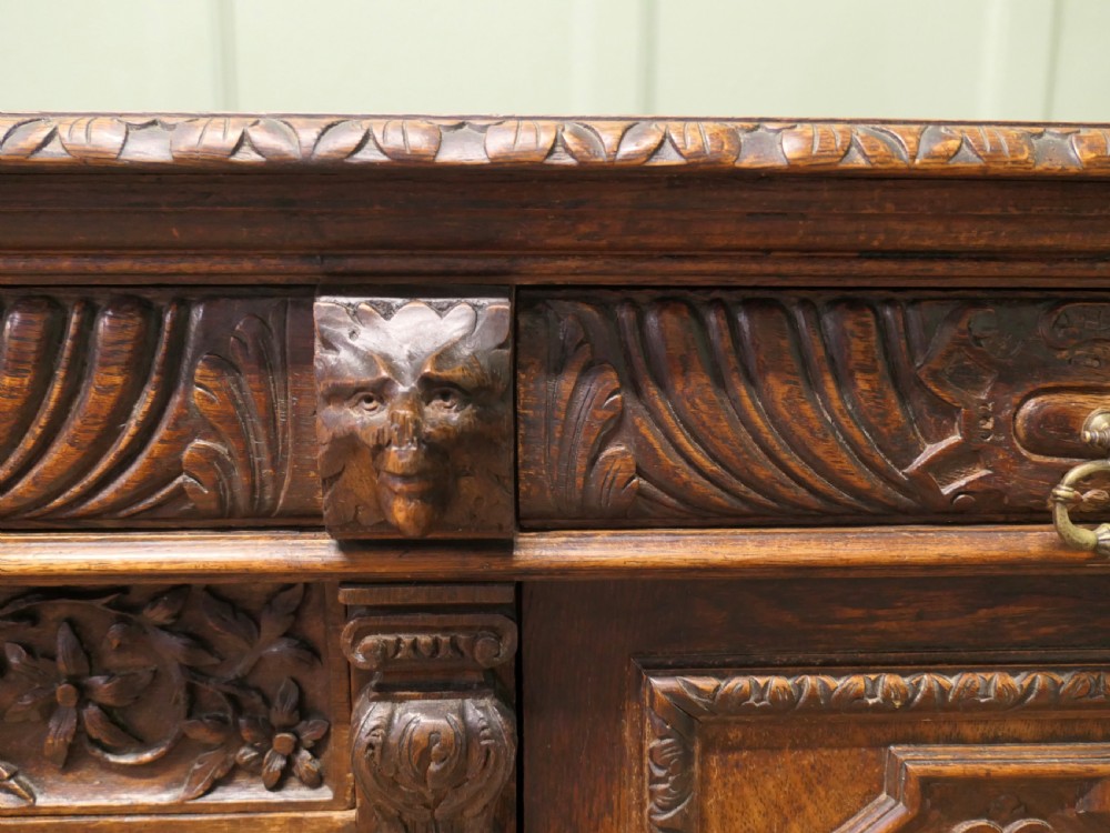A heavily carved warm brown oak sideboard with three top drawers, relief carved cupboard doors, putti side supports, and a central open recess. View III