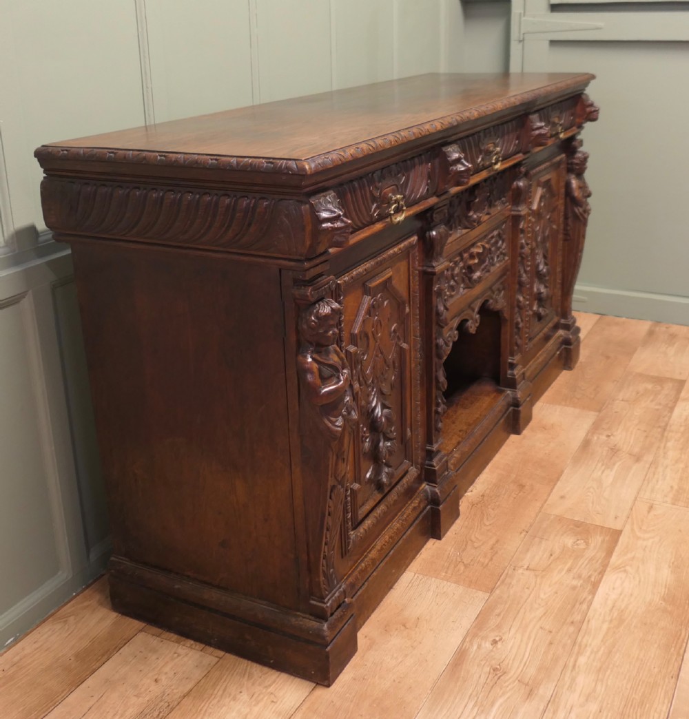 A heavily carved warm brown oak sideboard with three top drawers, relief carved cupboard doors, putti side supports, and a central open recess. View IV