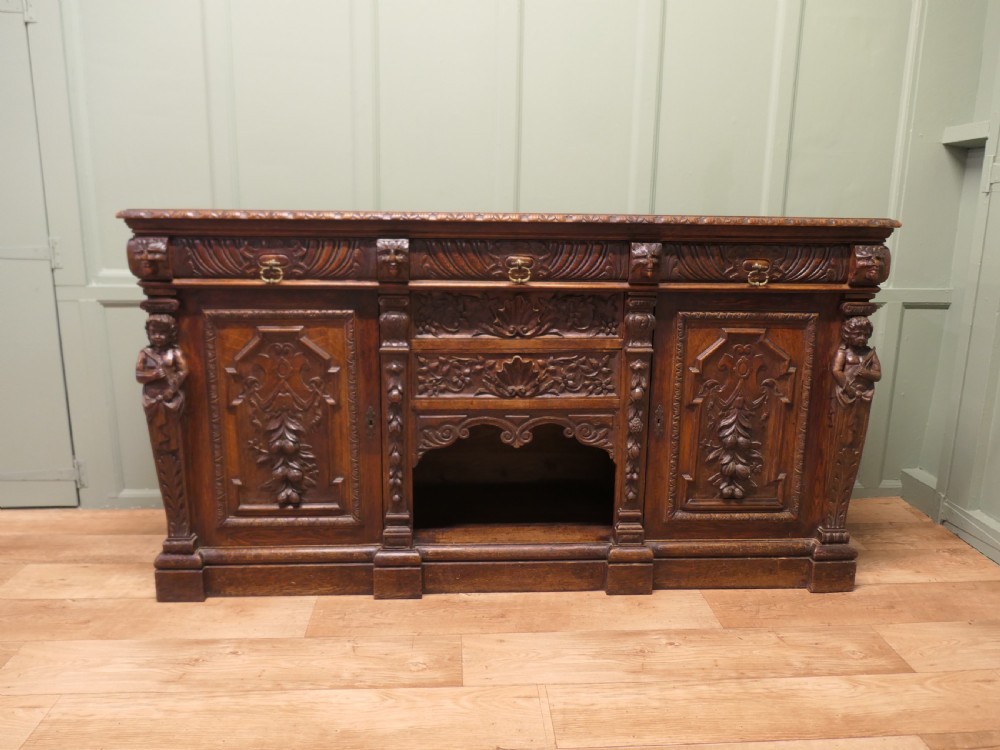 A heavily carved warm brown oak sideboard with three top drawers, relief carved cupboard doors, putti side supports, and a central open recess. View V