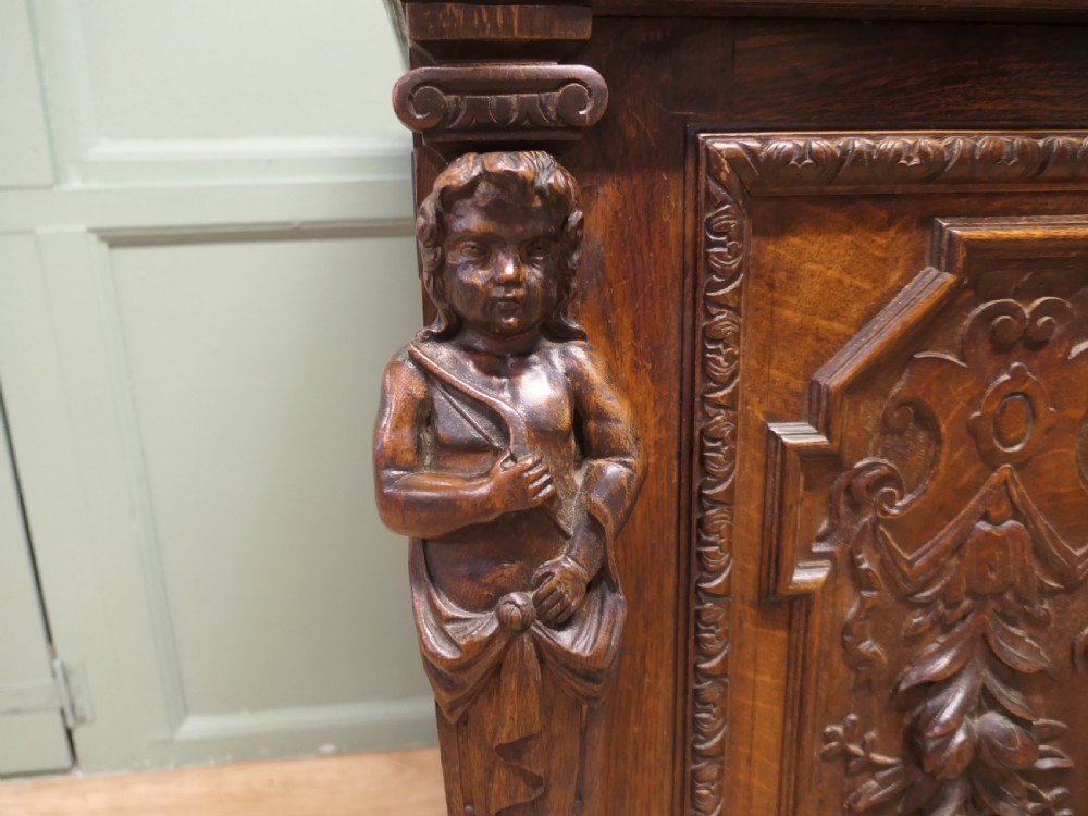 A heavily carved warm brown oak sideboard with three top drawers, relief carved cupboard doors, putti side supports, and a central open recess. View VI
