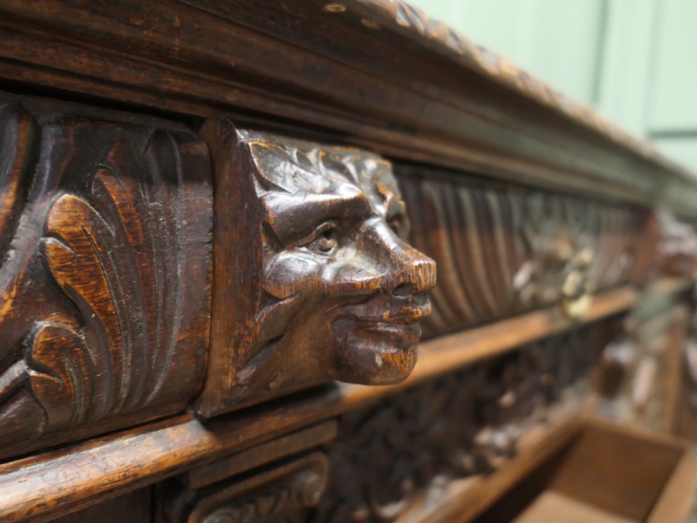 A heavily carved warm brown oak sideboard with three top drawers, relief carved cupboard doors, putti side supports, and a central open recess. View VII