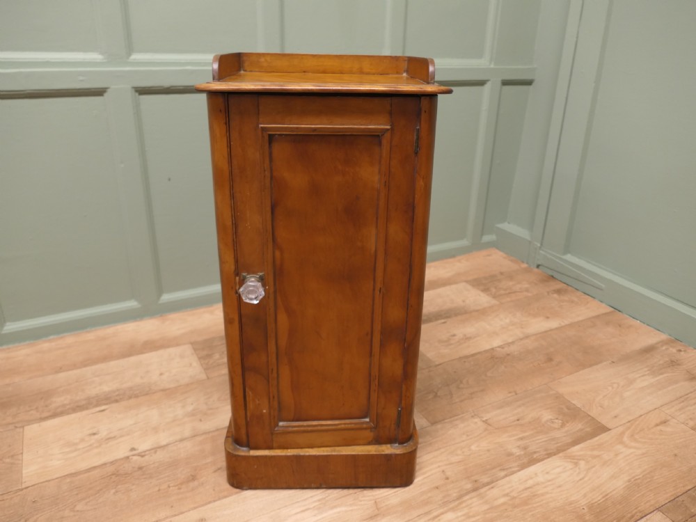 A Victorian birch bedside cupboard featuring a three-sided gallery top, a single panelled door with a glass handle, and a solid plinth base. View V