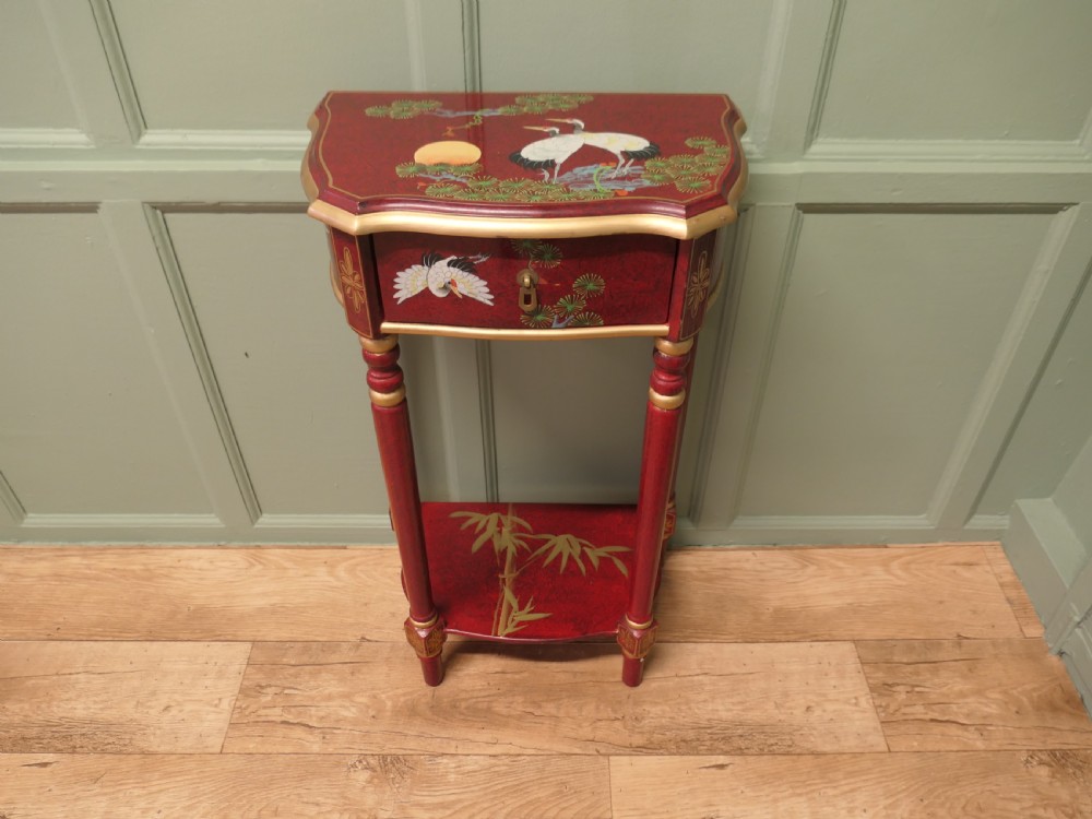 A tall red lacquer Chinoiserie side table featuring hand-painted crane and floral motifs, gilded turned legs, a single drawer, and an undershelf. View V