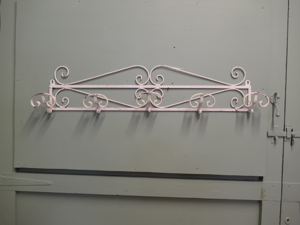 A long, wall-mounted off-white wrought iron hat and coat rack featuring scrollwork detailing and multiple curved hooks.