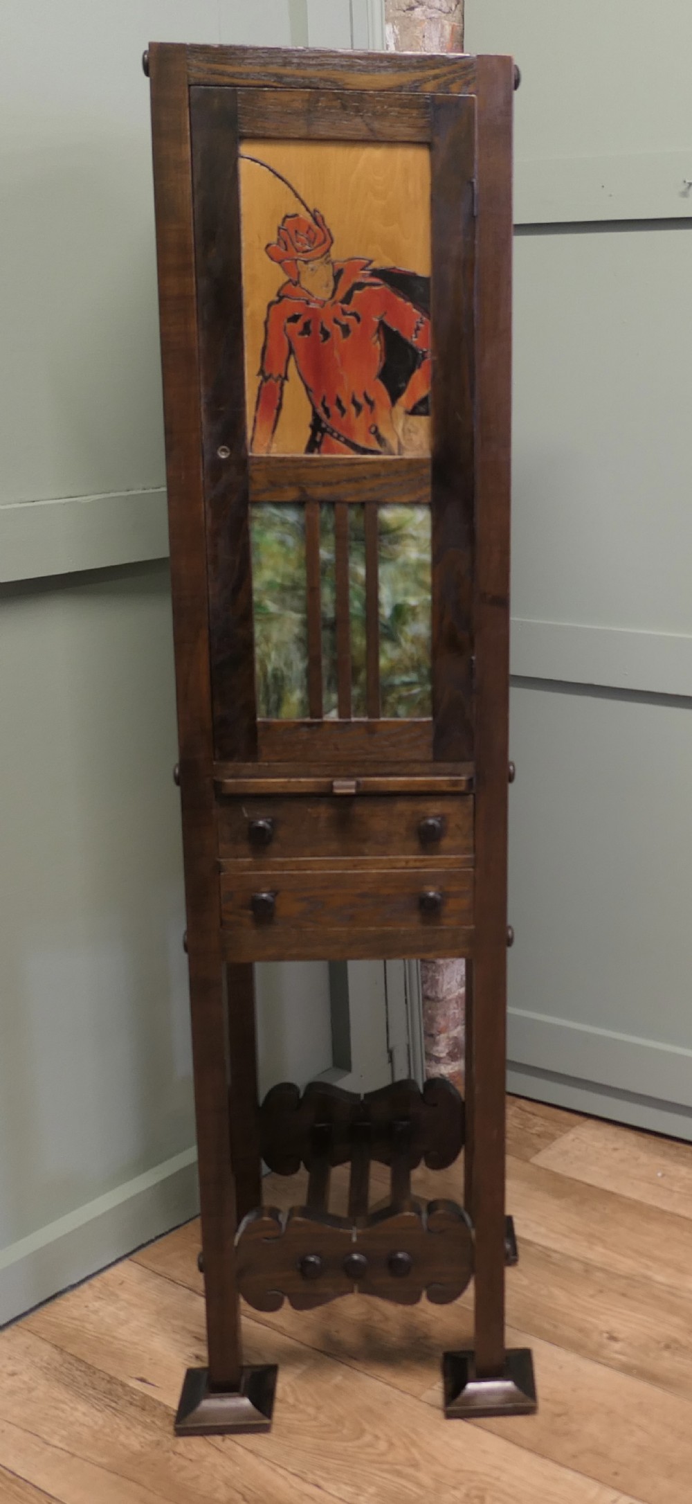 A tall, slender dark oak Arts and Crafts cabinet featuring a decorative inlaid wood panel depicting a figure in red, a slatted glass door section, two small drawers, and a lower shelf.
