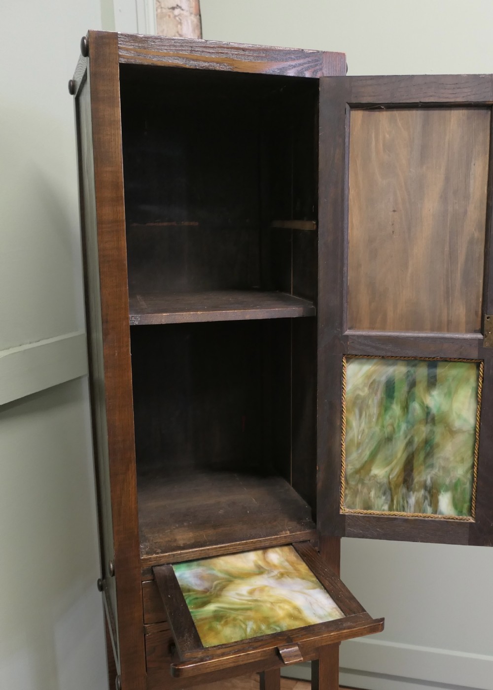 A tall, slender dark oak Arts and Crafts cabinet featuring a decorative inlaid wood panel depicting a figure in red, a slatted glass door section, two small drawers, and a lower shelf. View III