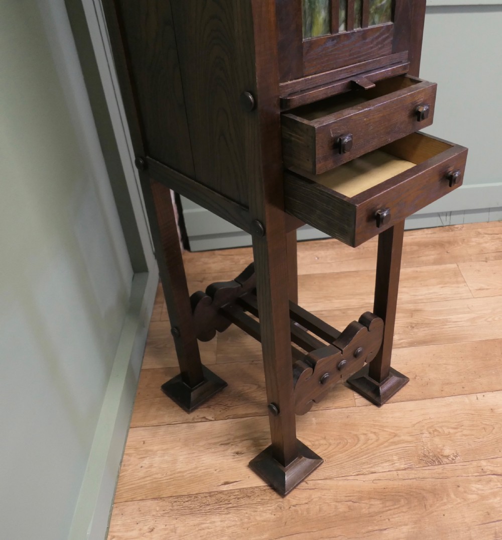 A tall, slender dark oak Arts and Crafts cabinet featuring a decorative inlaid wood panel depicting a figure in red, a slatted glass door section, two small drawers, and a lower shelf. View V