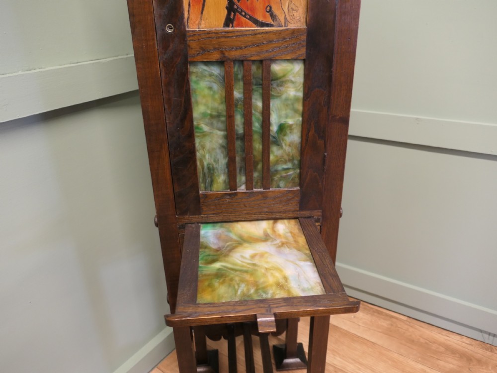 A tall, slender dark oak Arts and Crafts cabinet featuring a decorative inlaid wood panel depicting a figure in red, a slatted glass door section, two small drawers, and a lower shelf. View VI