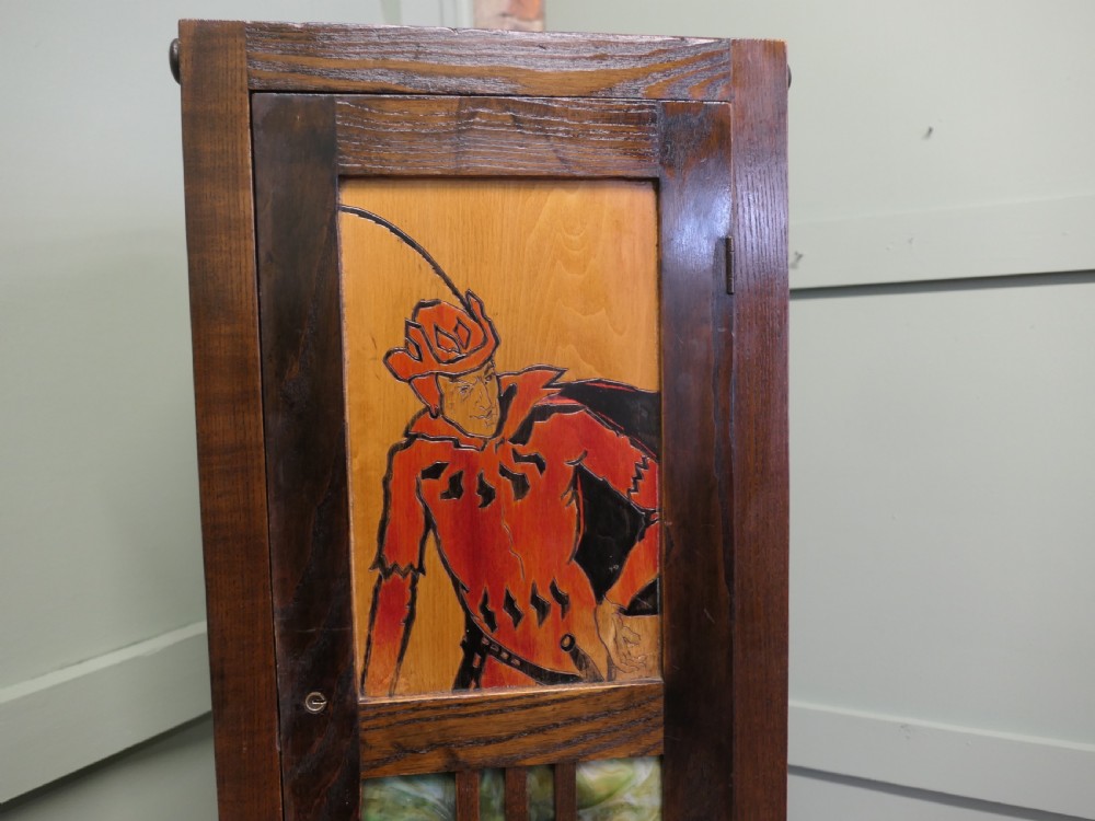 A tall, slender dark oak Arts and Crafts cabinet featuring a decorative inlaid wood panel depicting a figure in red, a slatted glass door section, two small drawers, and a lower shelf. View VII