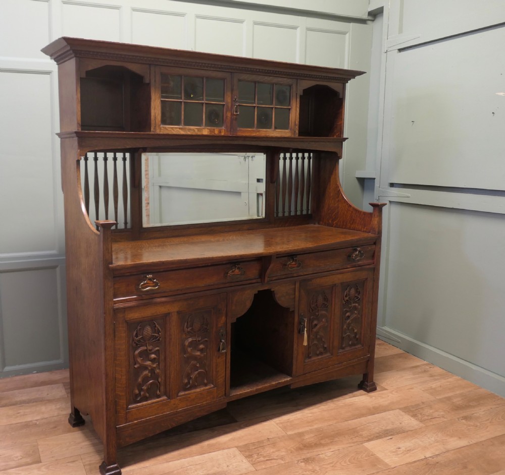 A large Arts and Crafts oak sideboard featuring a mirrored back, upper leaded glass cabinets, carved lower cupboard doors with botanical motifs, and brass handles.