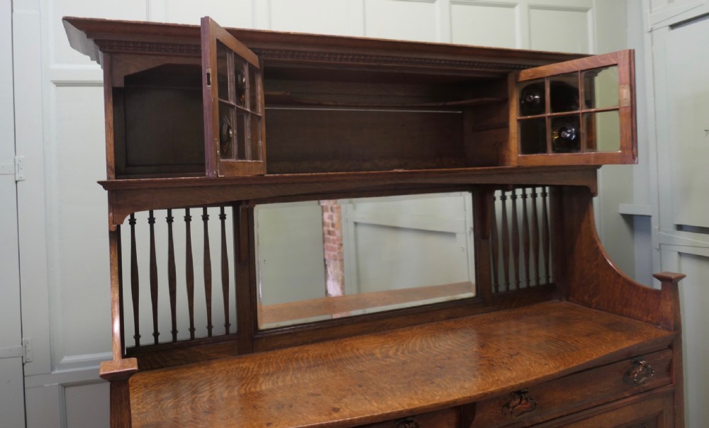 A large Arts and Crafts oak sideboard featuring a mirrored back, upper leaded glass cabinets, carved lower cupboard doors with botanical motifs, and brass handles. View II