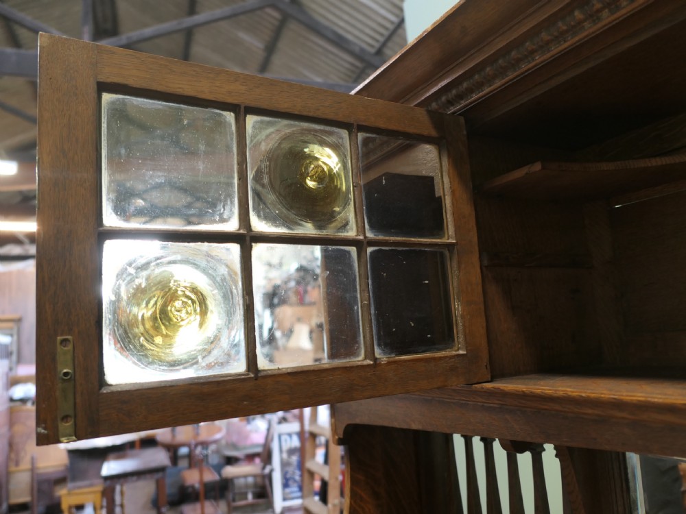 A large Arts and Crafts oak sideboard featuring a mirrored back, upper leaded glass cabinets, carved lower cupboard doors with botanical motifs, and brass handles. View III