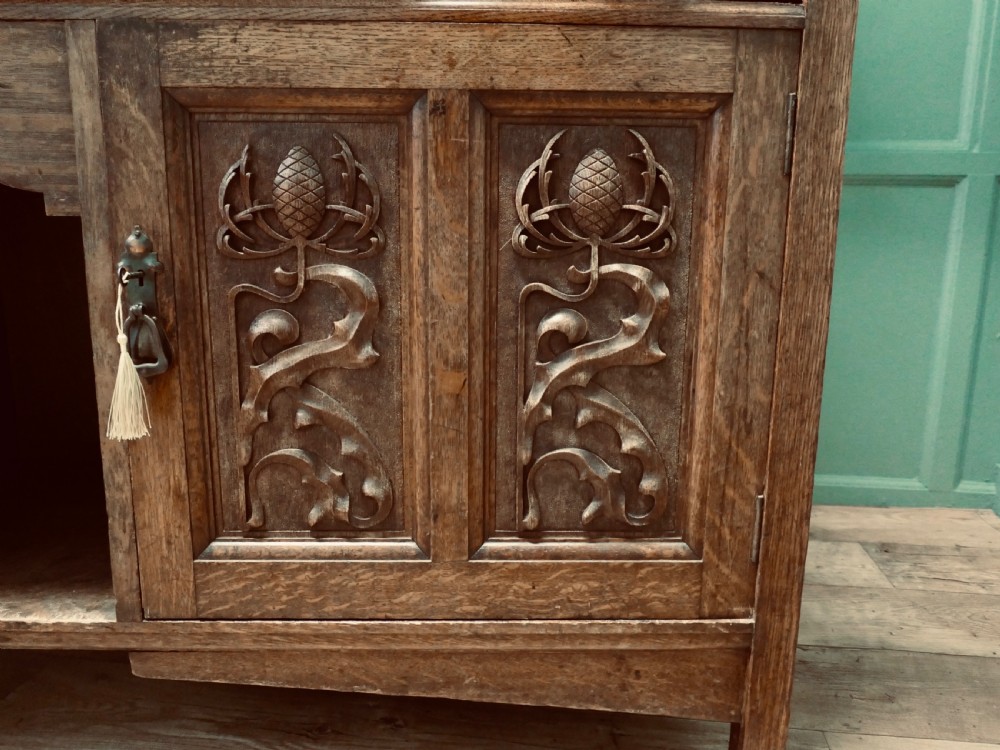 A large Arts and Crafts oak sideboard featuring a mirrored back, upper leaded glass cabinets, carved lower cupboard doors with botanical motifs, and brass handles. View VI