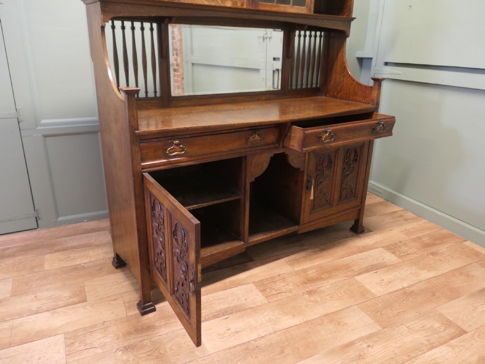 A large Arts and Crafts oak sideboard featuring a mirrored back, upper leaded glass cabinets, carved lower cupboard doors with botanical motifs, and brass handles. View VII