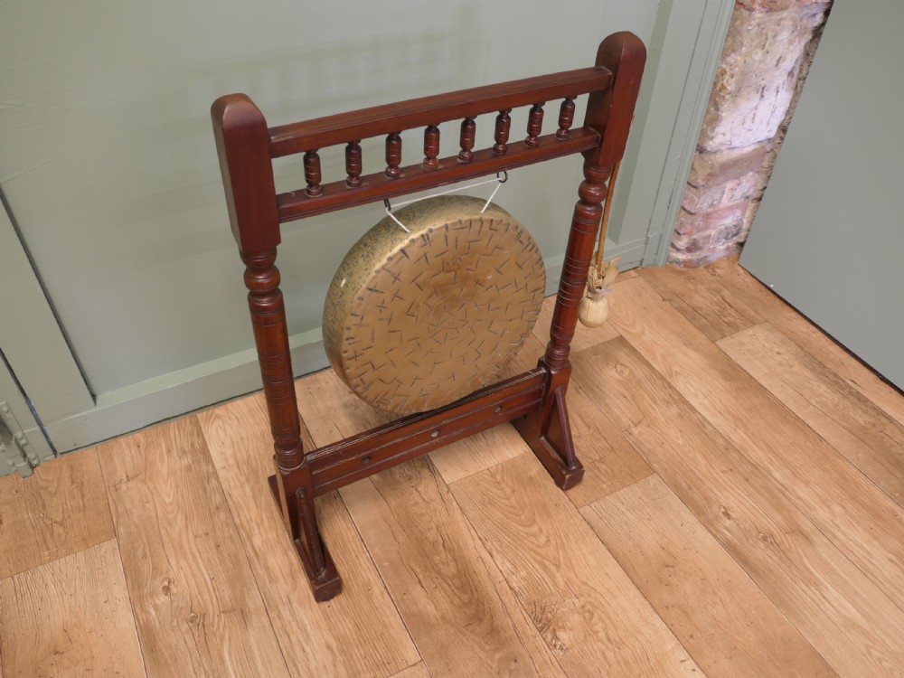 An Arts and Crafts dark timber floor frame featuring turned upper spindles and holding a central suspended hammered brass circular gong disc with an attached striker. View II