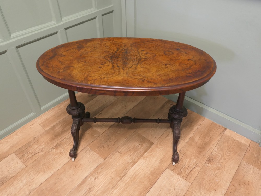 An oval Victorian centre table featuring a burr walnut top, turned stretcher base, carved legs, and white ceramic castors.