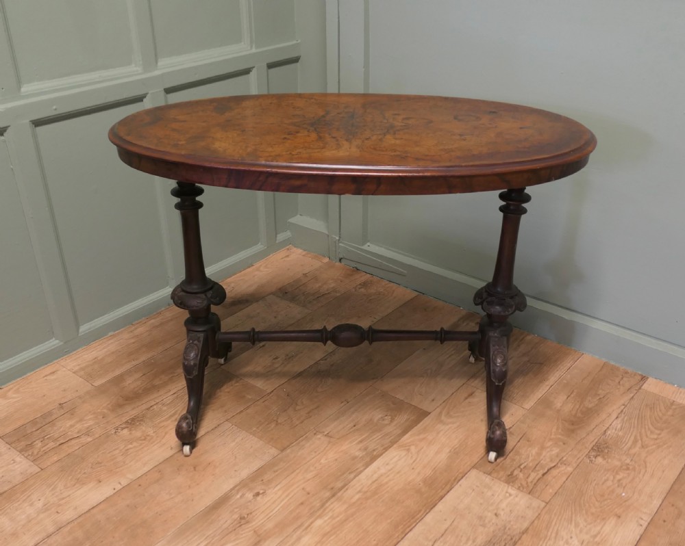 An oval Victorian centre table featuring a burr walnut top, turned stretcher base, carved legs, and white ceramic castors. View II