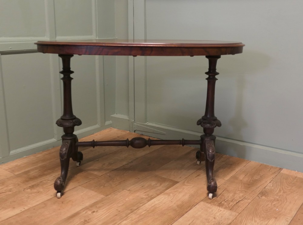 An oval Victorian centre table featuring a burr walnut top, turned stretcher base, carved legs, and white ceramic castors. View V