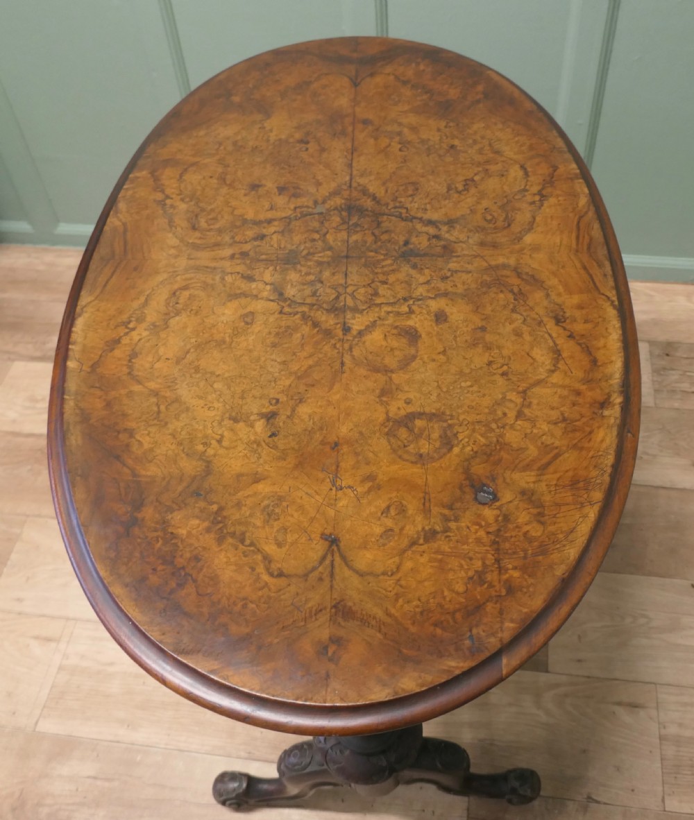 An oval Victorian centre table featuring a burr walnut top, turned stretcher base, carved legs, and white ceramic castors. View VI