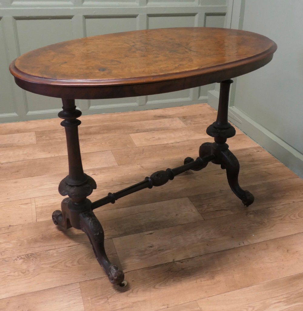 An oval Victorian centre table featuring a burr walnut top, turned stretcher base, carved legs, and white ceramic castors. View VII