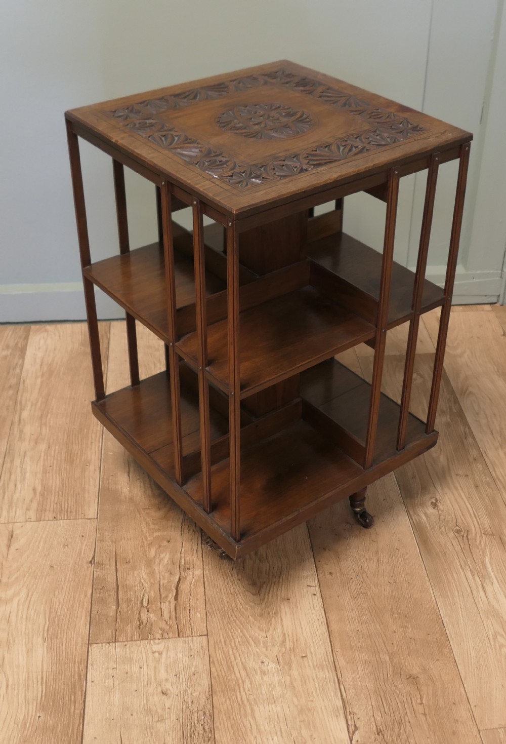 A square dark oak two-tier revolving bookcase with a carved rosette top panel, slender vertical side slats, and castors.
