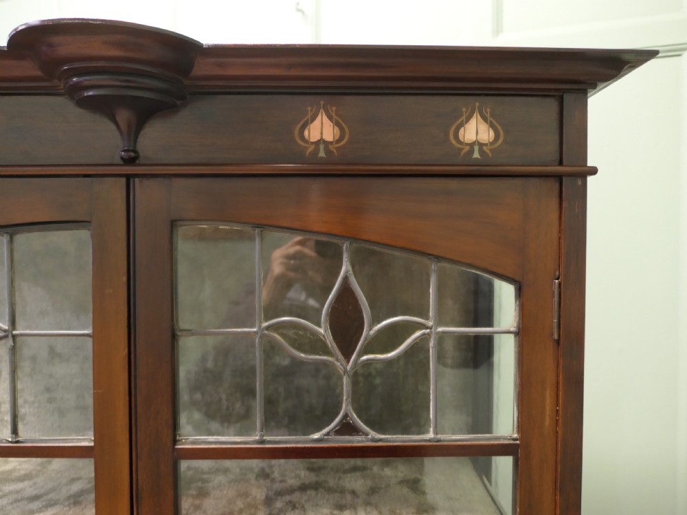 A tall Arts and Crafts mahogany display cabinet with leaded glass panels, slender flared legs, and floral wood inlay at the top. View V