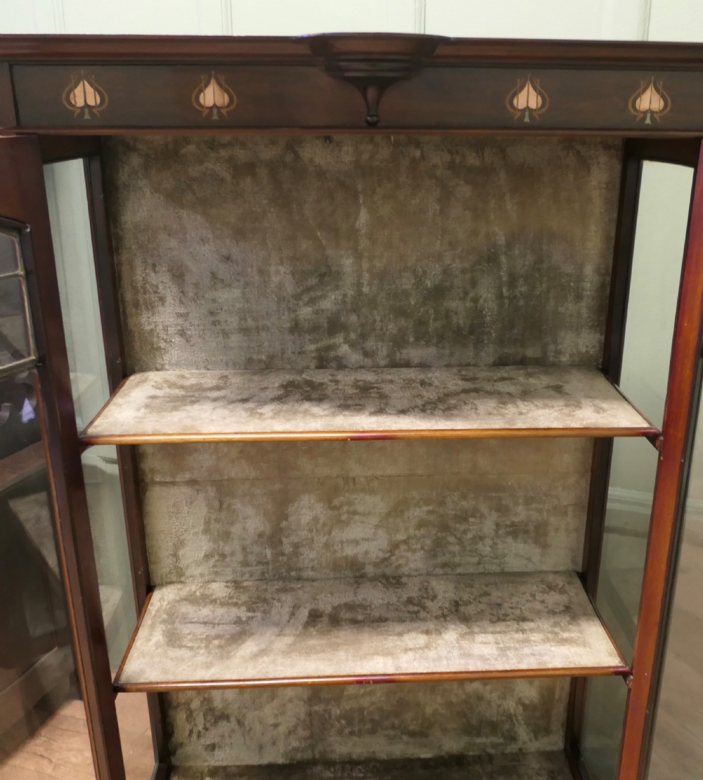 A tall Arts and Crafts mahogany display cabinet with leaded glass panels, slender flared legs, and floral wood inlay at the top. View VI