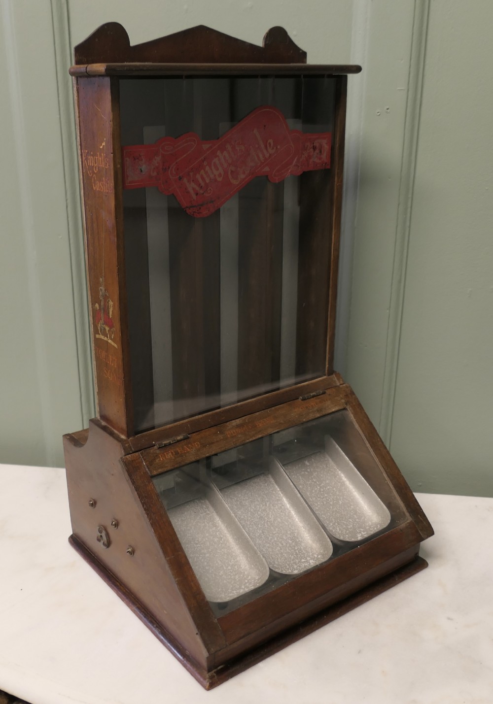 An antique wooden countertop soap dispensing cabinet featuring glazed panels, red Knights Castile branding, and three metal lower dispensing compartments. View III