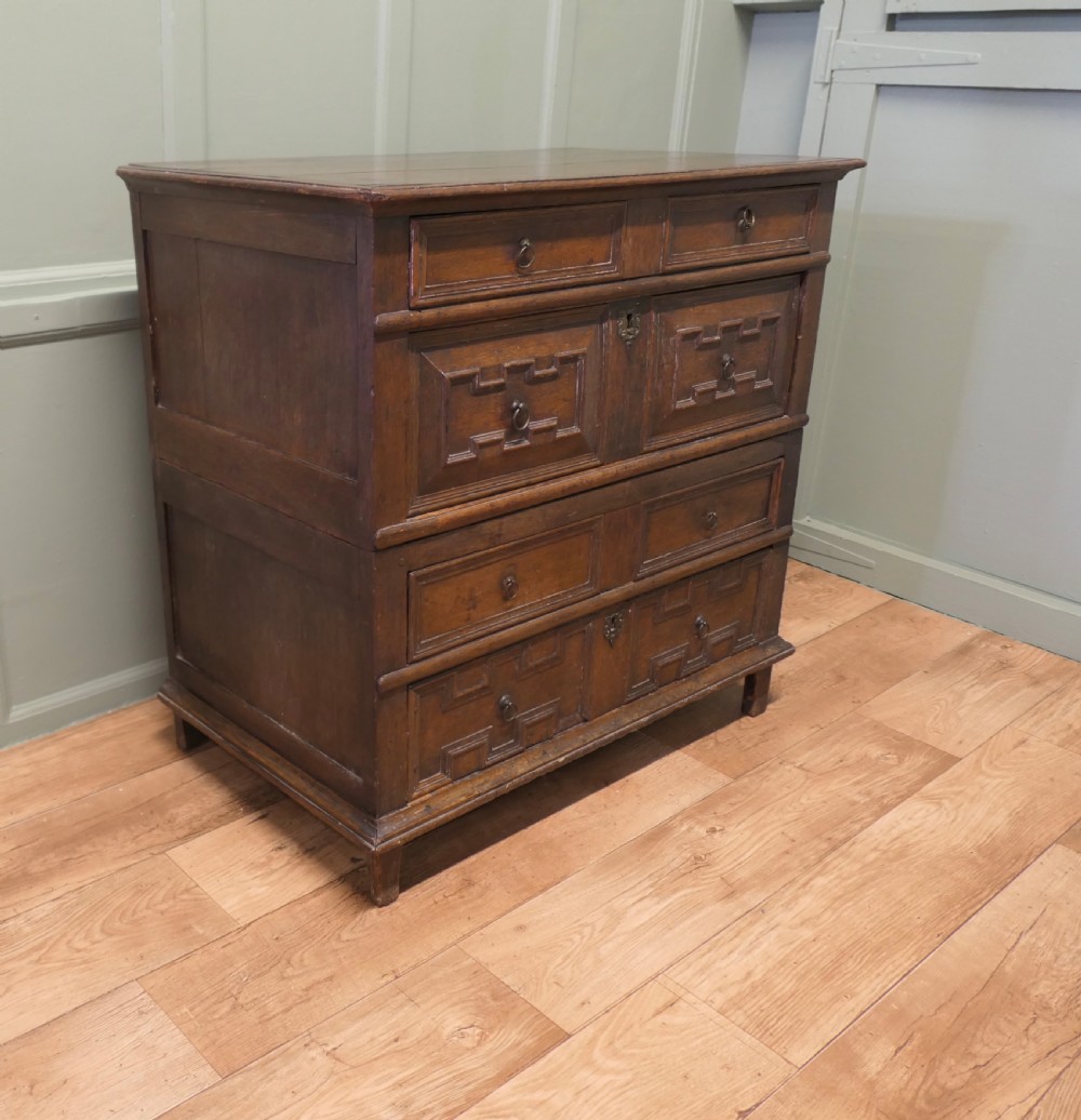An 18th century dark oak chest of drawers featuring geometric panelled drawer fronts, brass drop handles, and panelled sides.
