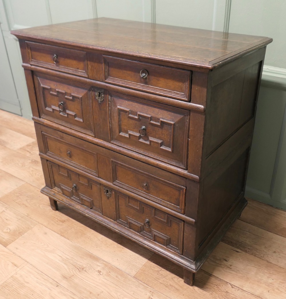 An 18th century dark oak chest of drawers featuring geometric panelled drawer fronts, brass drop handles, and panelled sides. View II
