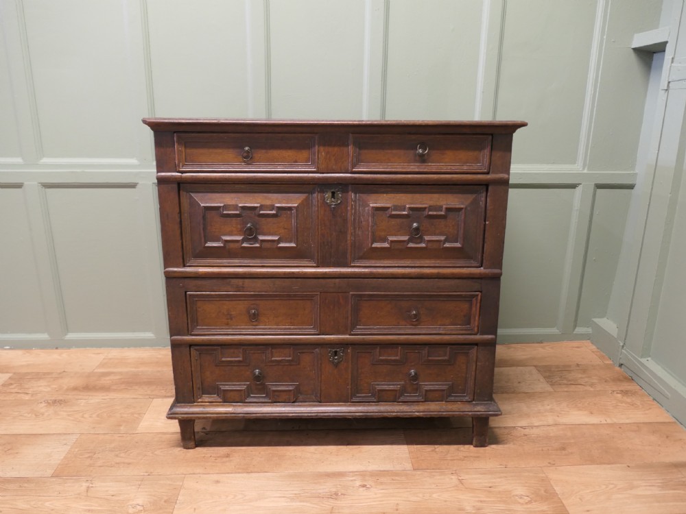 An 18th century dark oak chest of drawers featuring geometric panelled drawer fronts, brass drop handles, and panelled sides. View IV